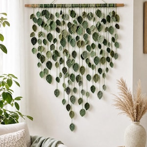 May include: A wall hanging featuring numerous crocheted green leaves suspended from a horizontal wooden dowel. The leaves vary in shades of green and are arranged in a cascading, V-shaped pattern, attached by thin green yarn strings to the dowel.