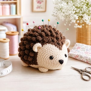 May include: A crochet hedgehog pincushion with a dark brown textured back and a cream-colored face. The hedgehog features black bead eyes, a small black nose, and several sewing pins with colorful round heads inserted into its back.