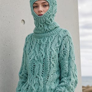 May include: A mint green cable-knit sweater featuring a matching integrated balaclava hood. The garment is crafted with a thick, textured yarn in a complex interwoven pattern, covering the head and neck while extending down to the torso.