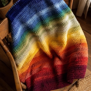 Könnte beinhalten: Eine gehäkelte Decke mit einem lebendigen Regenbogen-Farbverlauf, der durch Schattierungen von tiefem Blau, Grün, Gelb, Orange und Rot übergeht. Das Textil weist ein durchgehendes, strukturiertes Maschenmuster auf der gesamten Oberfläche auf.
