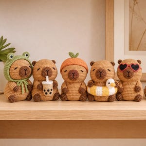 May include: Five small, tan, crocheted capybara figures arranged in a row. The first wears a green frog hood, the second holds a boba tea cup, the third wears an orange hat, the fourth has a yellow and white pool float, and the fifth wears heart-shaped sunglasses.