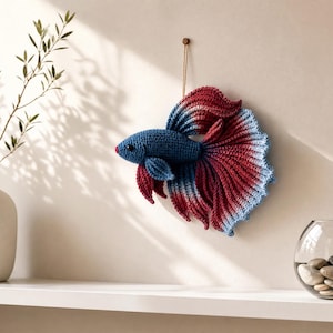 May include: A crocheted betta fish wall hanging featuring a gradient color pattern that transitions from deep blue at the head to vibrant red and light blue along the flowing, textured fins. The fish is suspended by a thin string against the wall.