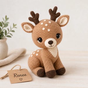 May include: A crocheted deer plush toy with light brown fur, white spots, and dark brown antlers. The deer has large black eyes and a cream-coloured muzzle. A brown paper tag attached by a string features the printed text Renee and a small heart symbol.