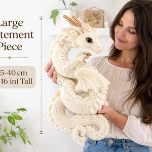 Puede incluir: Un dragón de peluche tejido a ganchillo en color crema con cuernos beige y ojos oscuros. El dragón presenta un cuerpo largo y enroscado con escamas texturizadas y alas. El texto indica Large Statement Piece y 35-40 cm de altura.