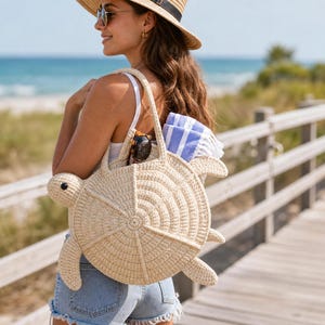 May include: A beige crochet tote bag shaped like a sea turtle with a circular shell body, a head, and four flippers. The bag features a textured, radial stitch pattern and two shoulder straps, with a blue and white striped towel tucked inside.