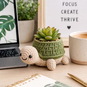 Può includere: Una statuetta di tartaruga all'uncinetto con testa e arti beige, dotata di un guscio verde scuro che funge da vaso per una piccola pianta succulenta verde. La tartaruga ha occhi a perla neri e piccole guance rosa ricamate.