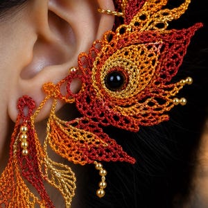 May include: An intricate ear cuff featuring a crochet design in shades of red, orange, and gold. The piece includes a central black bead, delicate thread-work patterns resembling leaves, and small gold-toned metallic beads attached to the edges.