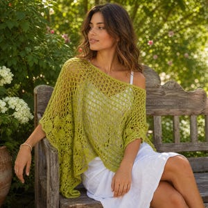 Op de afbeelding: Een limoengroene, grofgebreide gehaakte poncho met een delicaat, herhalend geometrisch patroon. Het kledingstuk heeft een brede, asymmetrische halslijn die over de schouders valt, gecombineerd met een eenvoudig wit mouwloos topje en een witte rok.