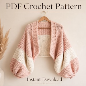 Peut inclure: Un modèle de crochet PDF pour un gilet court présentant un design color-block en rose poudré et crème. Le vêtement possède des manches larges et volumineuses ainsi qu'un point texturé, présenté sur un cintre en bois avec le texte PDF Crochet Pattern.
