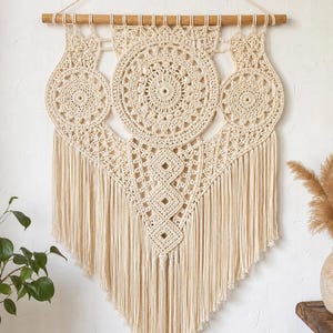 Könnte beinhalten: Ein cremefarbener Makramee-Wandbehang mit drei kreisförmigen Häkel-Mandalas, die an einem horizontalen Holzstab hängen. Das Design umfasst komplizierte Knotenmuster und eine lange, V-förmige Franse, die an der Unterkante herabhängt.