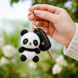 May include: A small, crocheted panda bear keychain featuring a white and black yarn body, round black ears, black eye patches, and pink stitched cheeks. The panda is attached to a gold-toned metal split ring and chain alongside a black car key.