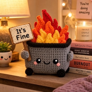 May include: A gray crocheted dumpster plushie with a smiling face, pink cheeks, and black eyes. It holds a small rectangular sign that says "It's Fine" in black text. Inside the dumpster are crocheted flames in shades of bright orange, red, and yellow.