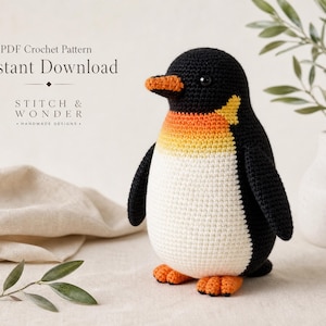 Op de afbeelding: Een gehaakt pinguïnspeeltje met een zwarte kop, rug en vleugels, een witte buik en een oranje snavel en voeten. De borst heeft een kleurverloop van oranje naar geel. De tekst luidt: PDF Crochet Pattern Instant Download STITCH & WONDER.