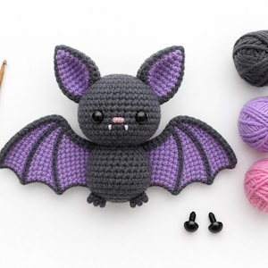 Può includere: Un pipistrello di peluche all'uncinetto grigio scuro e viola con occhi di sicurezza neri, un naso rosa e due zanne bianche. Accanto ci sono un uncinetto in legno da 2,5 mm, tre gomitoli di lana grigia, viola e rosa e due occhi di sicurezza neri.