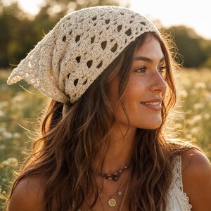 May include: A cream-coloured crochet headscarf featuring an intricate openwork pattern. The triangular-shaped accessory is tied securely at the back of the head, showcasing a delicate, textured knit design with a scalloped edge along the border.