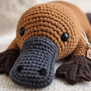 May include: A crocheted platypus toy with a light brown body, a dark grey bill, and dark brown webbed feet. The toy features black plastic eyes and a small wooden tag attached to the side with the name Hazel and a small heart symbol engraved.