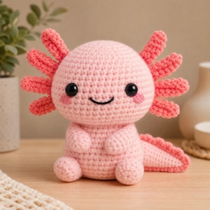 Könnte beinhalten: Ein hellrosa gehäkeltes Axolotl-Stofftier mit rundem Kopf, großen schwarzen Kunststoffaugen, einem kleinen gestickten Lächeln und rosa Wangen. Es hat drei federartige äußere Kiemen auf jeder Kopfseite und einen strukturierten Schwanz.
