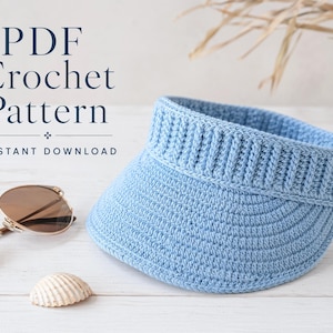 May include: A light blue crochet visor with a ribbed headband and a curved brim. The text "PDF Crochet Pattern" and "INSTANT DOWNLOAD" is printed in dark blue. A pair of brown-tinted sunglasses and a small white seashell are positioned nearby.