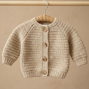 May include: A beige long-sleeved cardigan made of thick crochet yarn. The garment features a round neckline, ribbed cuffs, and a front placket secured with four round, light brown wooden buttons. It is displayed on a woven wicker hanger.