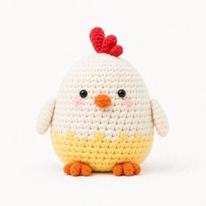 Puede incluir: Un juguete de peluche de pollo tejido a ganchillo de forma redonda, con cuerpo superior blanco y cuerpo inferior amarillo con borde dentado. Tiene una cresta roja brillante en la parte superior, un pico naranja, dos ojos de cuentas negras y mejillas rosadas.