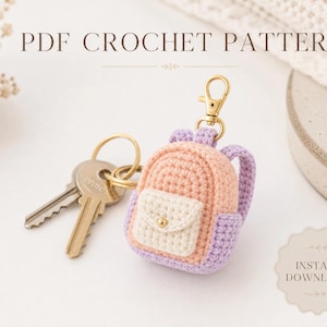 May include: A crochet backpack keychain in peach, lavender, and cream with a gold-tone lobster clasp and a metal key ring holding two silver keys. The image includes the text PDF CROCHET PATTERN and INSTANT DOWNLOAD in a decorative font.