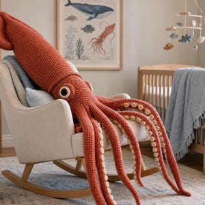 May include: A large, rust-colored crochet squid plush toy with a textured surface, prominent dark eyes, and multiple long tentacles featuring small, circular cream-colored suction cups, resting on a light-colored upholstered rocking chair.