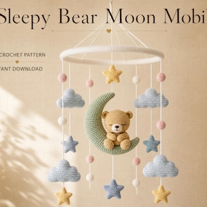 Può includere: Una giostrina all'uncinetto con un orsetto beige seduto su una mezzaluna verde salvia, circondato da nuvole azzurre, stelle gialle e blu e piccole sfere rosa e bianche appese a un cerchio bianco. Il testo recita Sleepy Bear Moon Mobile.