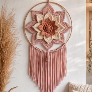 Peut inclure: Une décoration murale circulaire composée d'une fleur de lotus crochetée à plusieurs niveaux dans des tons rose poudré, crème et terre cuite. La fleur est fixée dans un cerceau en bois, avec de longues franges en macramé rose poudré.