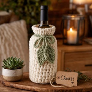 Peut inclure: Une housse de bouteille crochetée de couleur crème avec un motif de points texturés, un nœud décoratif crocheté vert et deux feuilles crochetées vertes fixées sur le devant. Une étiquette cadeau en papier brun avec le texte Cheers! et un cœur.