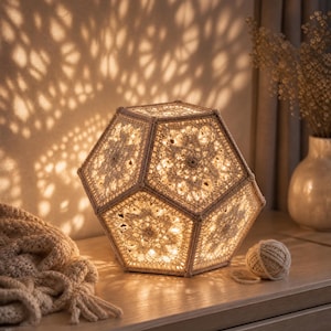 Op de afbeelding: Een dodecaëdervormige lamp opgebouwd uit beige gehaakte panelen met ingewikkelde bloemmotieven. De lamp verspreidt een warm, gloeiend licht dat gedetailleerde, geometrische schaduwpatronen op het omliggende wandoppervlak werpt.