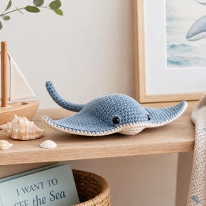 May include: A blue and cream-colored crocheted stingray plush toy with black safety eyes. The stingray features a textured yarn surface, a wide body with pointed wings, and a long, slender tail extending from the back of the main body.