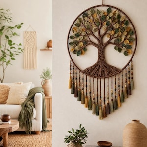 Puede incluir: Un tapiz circular para pared que presenta un diseño de árbol de la vida en macramé marrón con hojas de tela verdes y amarillas. La pieza está enmarcada por un aro marrón e incluye hilos colgantes decorados con cuentas de madera y borlas.