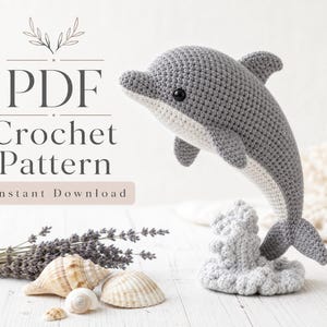 May include: A gray and white crocheted dolphin toy standing on a crocheted wave base. The text PDF Crochet Pattern Instant Download is visible on the left side. The dolphin has a dark eye and a smooth, textured yarn surface throughout its body.