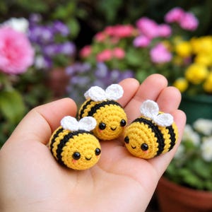Op de afbeelding: Drie kleine, ronde, gehaakte bijenfiguren met gele en zwarte horizontale strepen, witte vleugels en zwarte kraaloogjes. Elke bij heeft een glimlachende uitdrukking met kleine roze blosjes op de wangen, gemaakt van garen.