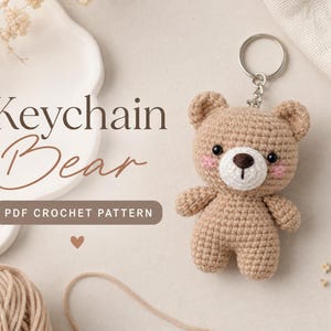May include: A light brown crocheted bear keychain with a white muzzle, dark eyes, and pink cheeks. The text reads Keychain Bear PDF CROCHET PATTERN. The bear features a metal ring and chain attached to the top of its head for hanging purposes.