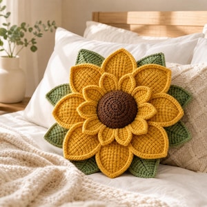 May include: A decorative sunflower-shaped throw pillow featuring a dark brown circular center, multiple layers of bright yellow crocheted petals, and green crocheted leaves extending from the base, all crafted with a textured yarn finish.