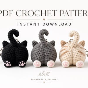 May include: Three crochet cat butt keychains in black, gray, and beige with pink paw pads and upright tails. The text PDF CROCHET PATTERN INSTANT DOWNLOAD and HANDMADE WITH LOVE appears above and below the items on a plain white background.
