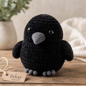 Può includere: Un uccellino di peluche lavorato all'uncinetto di colore nero con una finitura in filato testurizzato, dotato di un piccolo becco grigio, occhi in plastica scura e zampe grigie. Un'etichetta rettangolare marrone chiaro riporta il nome Oskar.