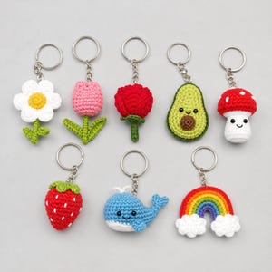 Puede incluir: Una colección de ocho llaveros de ganchillo con diversas formas, incluyendo una flor blanca, un tulipán rosa, una rosa roja, un aguacate verde, una seta roja y blanca, una fresa roja, una ballena azul y un arcoíris colorido con nubes.