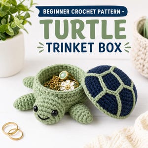 Può includere: Un portagioie a forma di tartaruga all'uncinetto con corpo verde chiaro e guscio blu scuro e verde chiaro. Il coperchio è rimosso, rivelando una catenina dorata e un anello a forma di fiore. Testo: BEGINNER CROCHET PATTERN - TURTLE TRINKET BOX.