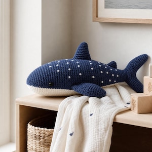 May include: A dark blue crocheted whale shark plush toy featuring a white underbelly and small white polka dot patterns on its back, with a prominent dorsal fin, side fins, and a forked tail, crafted with a textured yarn finish.