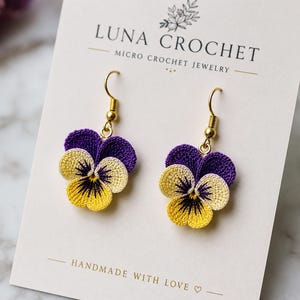 May include: A pair of micro crochet pansy flower earrings featuring purple, cream, and yellow thread with dark centre details. The earrings hang from gold-toned metal hooks and are displayed on a white card labelled LUNA CROCHET.
