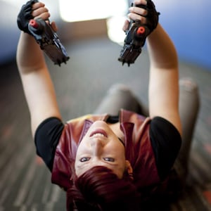 May include: A person with short reddish-brown hair wearing a black short-sleeved shirt and a dark red vest, holding two black toy pistols with orange tips in black fingerless gloves, viewed from an overhead perspective while lying down.