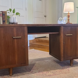 May include: A rectangular wooden desk featuring a dark brown finish, two side cabinets with single doors, and horizontal metal handles. The desk has a central open knee space and is supported by short, tapered legs. The surface is clear of items.
