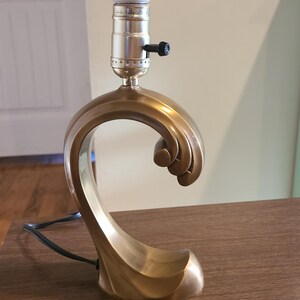 May include: A table lamp with a gold-colored, curved, wave-like base. The lamp features a metallic finish and a central vertical socket holding a single white light bulb. A small black knob is attached to the side of the socket for operation.