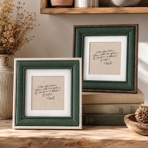 May include: Two square picture frames with dark green fabric borders and white inner mats. Each frame contains a beige insert with black handwritten text that reads, I love you more than all the grains of sand on a beach! with a red heart and Mom & Dad.