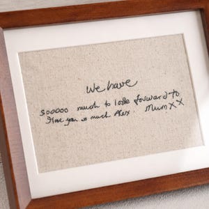 Puede incluir: Un marco de madera rectangular con un paspartú blanco que rodea una tela de color beige claro. La tela presenta un texto bordado en negro con letra cursiva que dice: We have sooooo much to look forward to. Love you so much Alex. Mum xx.