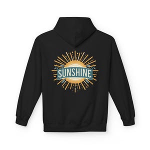 Puede incluir: Una sudadera negra con cremallera y capucha que presenta un gráfico en la espalda. El diseño muestra un sol amarillo estilizado con rayos y una banda azul en el centro que contiene el texto blanco SUNSHINE en una tipografía sans-serif.