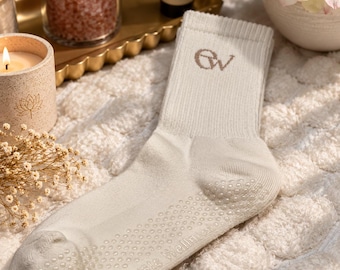 May include: A cream-colored ribbed crew sock featuring the embroidered letters EW on the cuff and the text Elevate Wellness printed in small white silicone grip dots on the sole, designed with a non-slip textured pattern on the bottom.