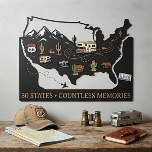 Könnte beinhalten: Eine schwarze Metall-Wandkarte der Vereinigten Staaten mit weißen Bergsilhouetten, einer kurvenreichen Straße und verschiedenen Reise-Symbolen wie einem Wohnmobil, einer Kamera und Kakteen. Der Text 50 STATES • COUNTLESS MEMORIES ist unten in Weiß aufgedruckt.